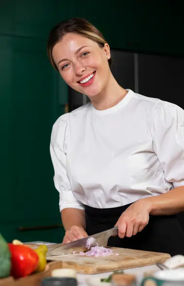 MY-PIC-2 Professional chef Chef Lina standing in a cozy kitchen, smiling while preparing a dish, surrounded by fresh ingredients and cooking tools.
