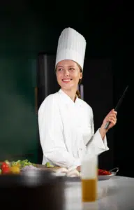 Professional chef Chef Lina standing in a cozy kitchen, smiling while preparing a dish, surrounded by fresh ingredients and cooking tools.